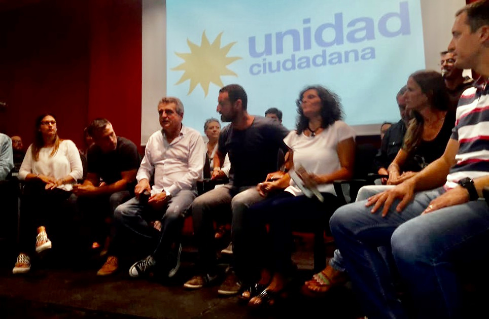 Participantes sentados en un escenario, con una proyección de Unidad Ciudadana al fondo.