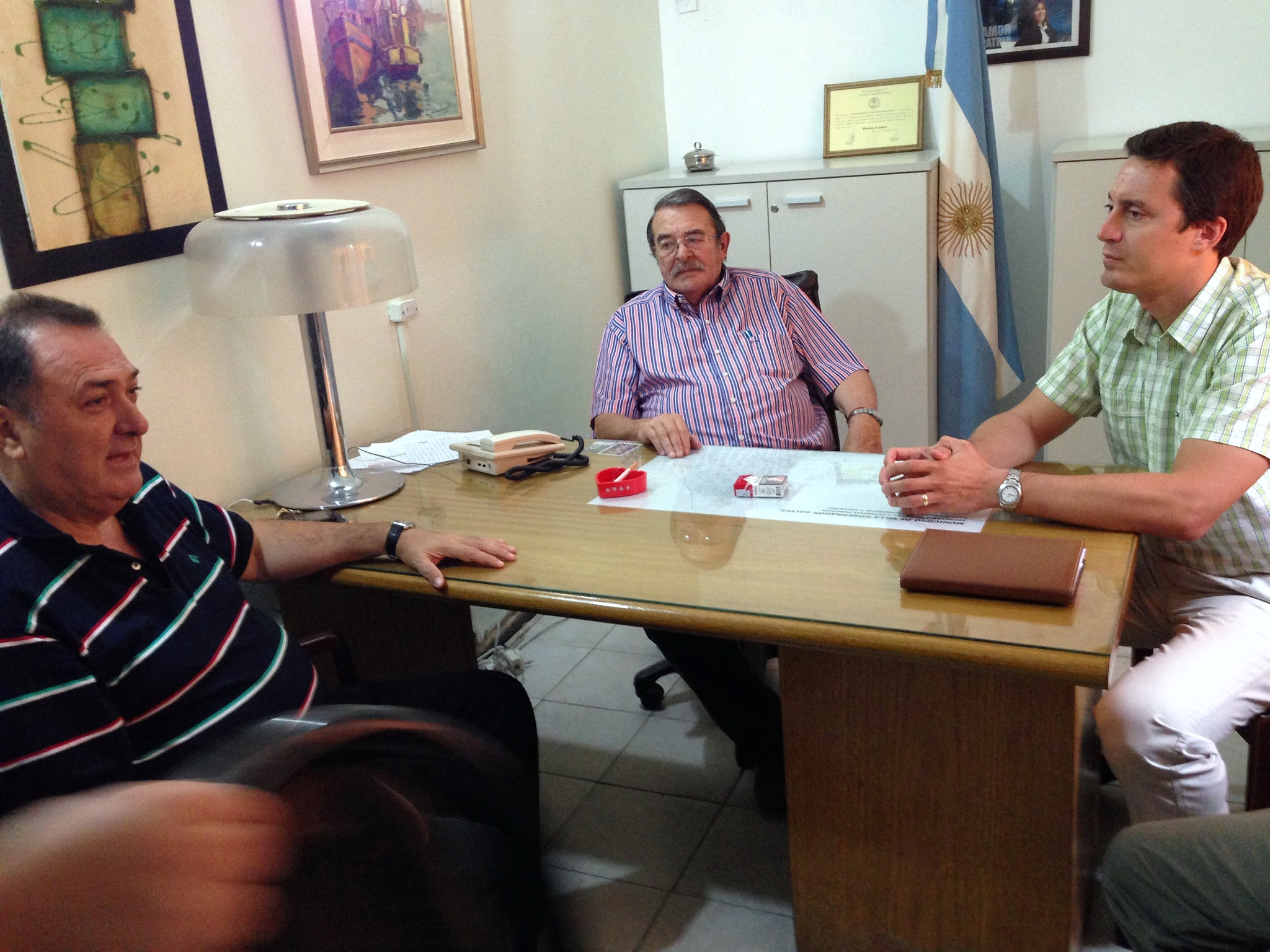 Tres personas reunidas alrededor de un escritorio, con una bandera argentina al fondo.