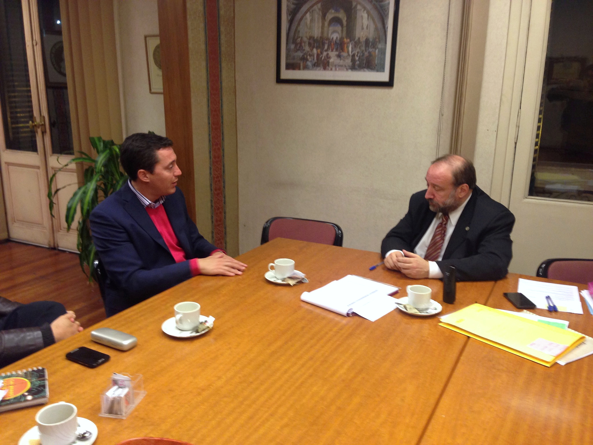 Dos personas conversan a ambos lados de una mesa con documentación y tazas.