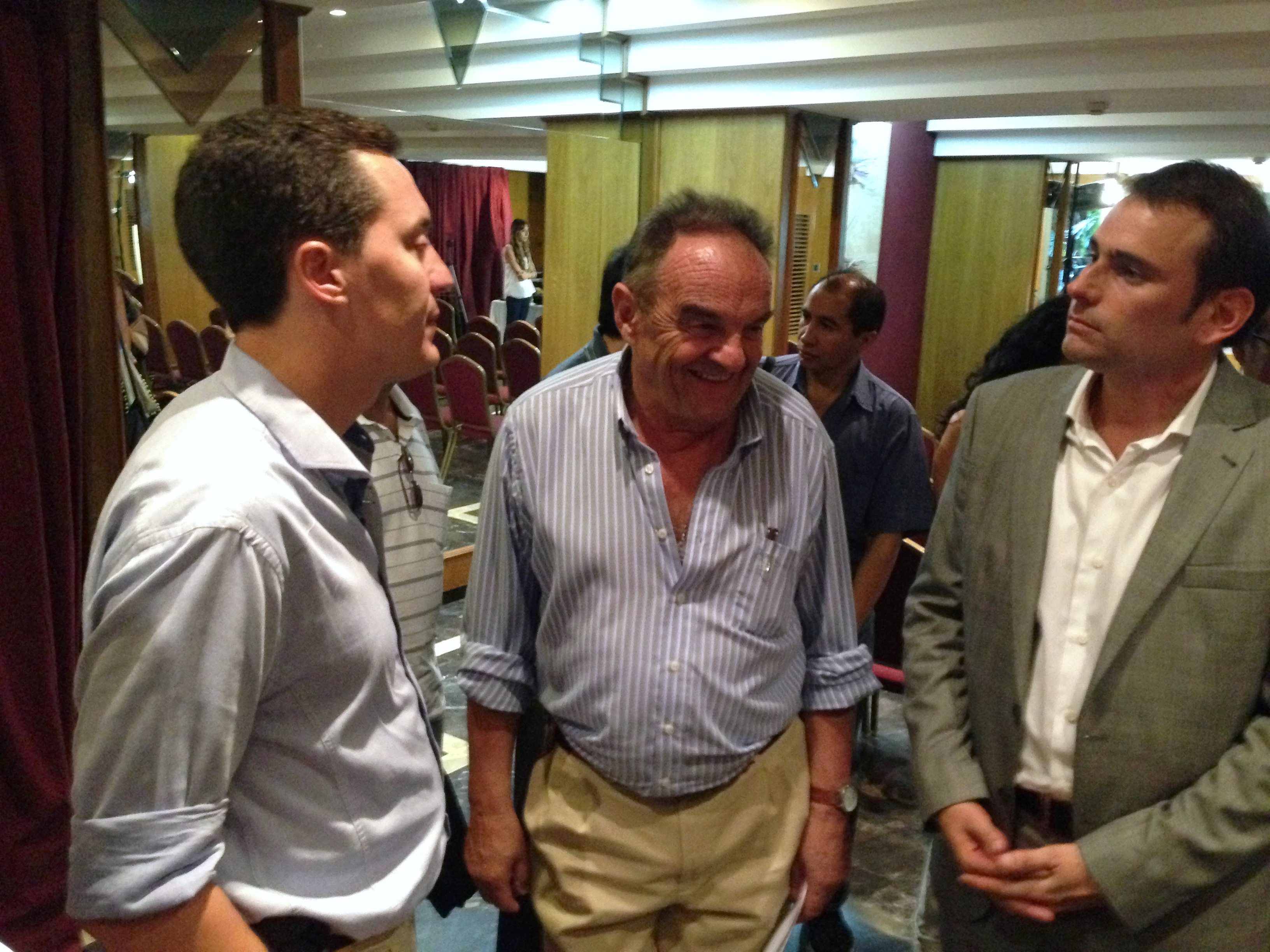 Tres personas en primer plano conversan en un salón con otras personas al fondo.