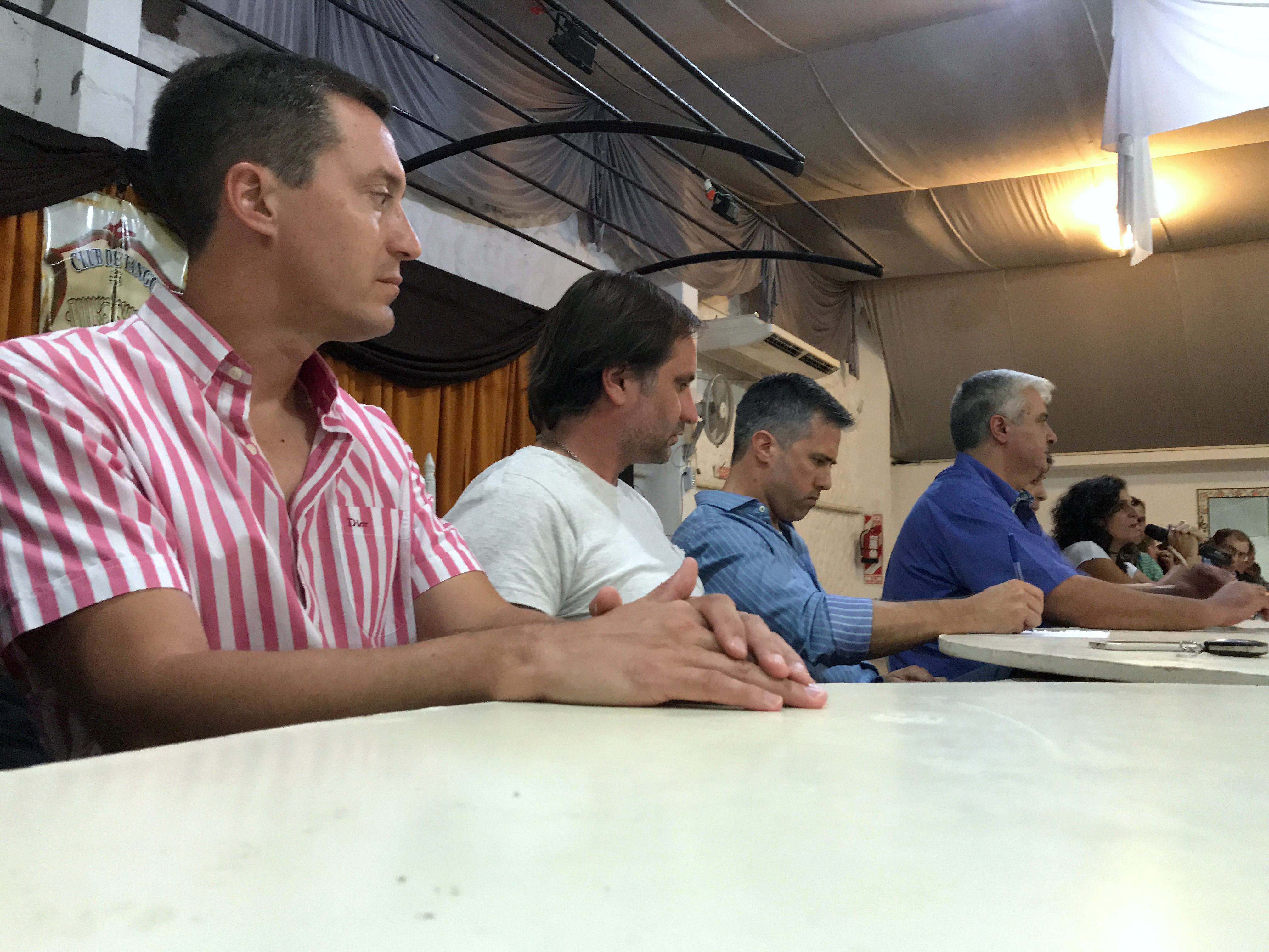 Personas sentadas en una mesa durante una reunión; al fondo una participante habla con un micrófono.