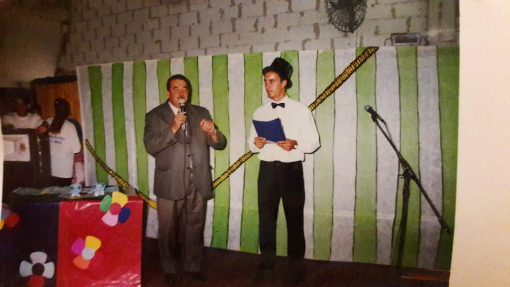 Dos personas en un escenario; una habla con micrófono y otra sostiene una carpeta.