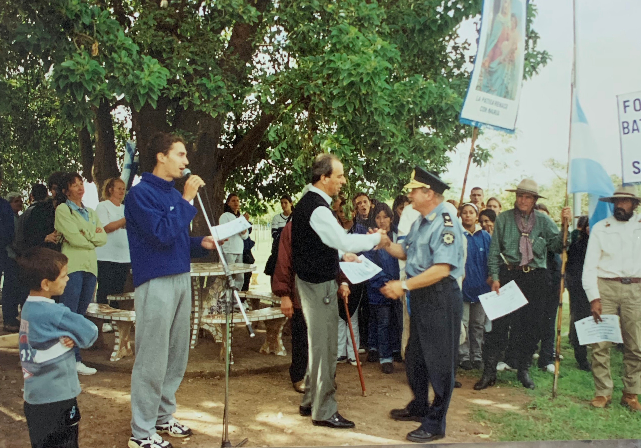 Acto al aire libre con participantes, banderas y una persona hablando al micrófono.
