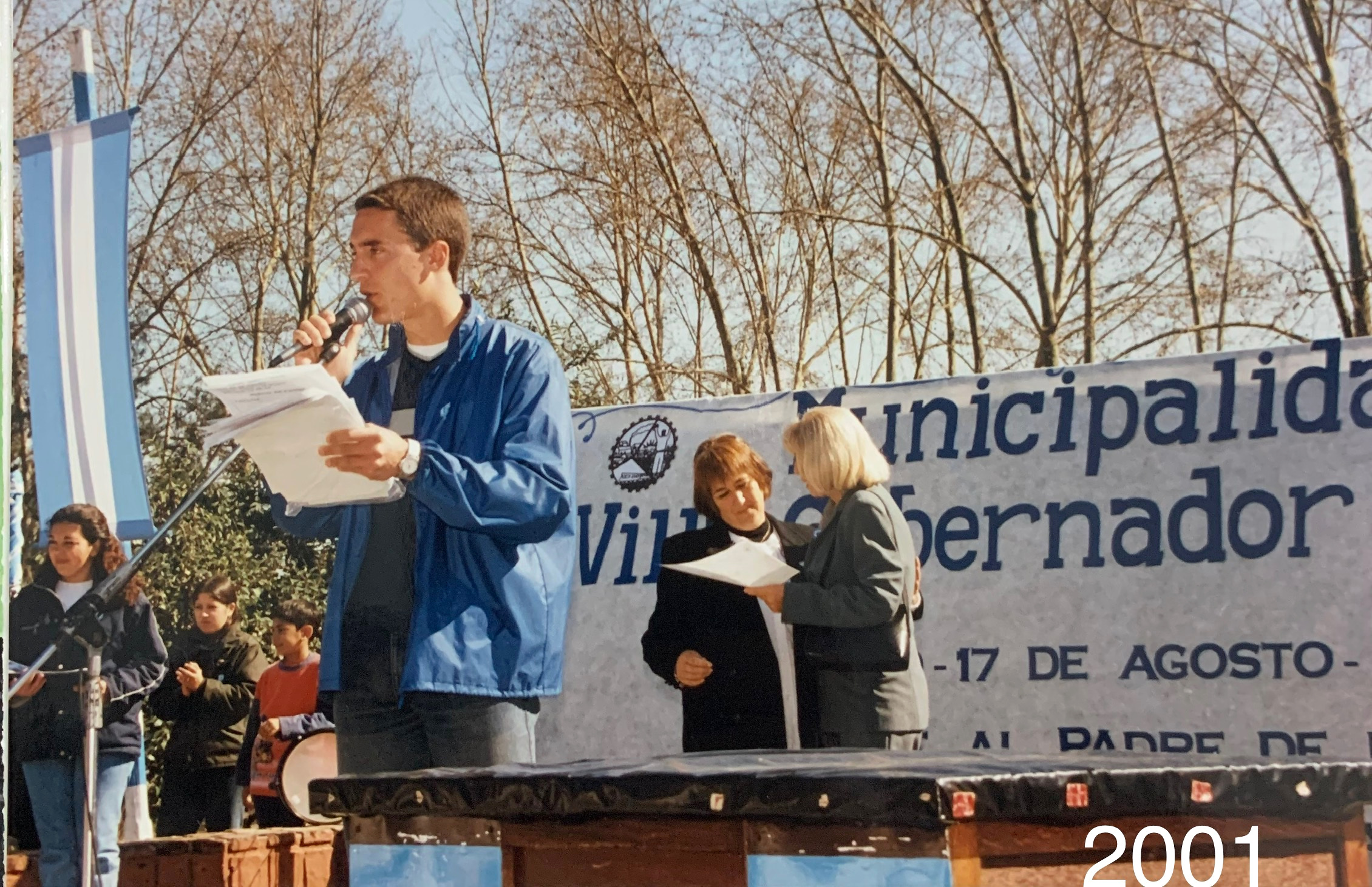Una persona habla al micrófono durante un acto al aire libre; la fotografía lleva la inscripción 2001.