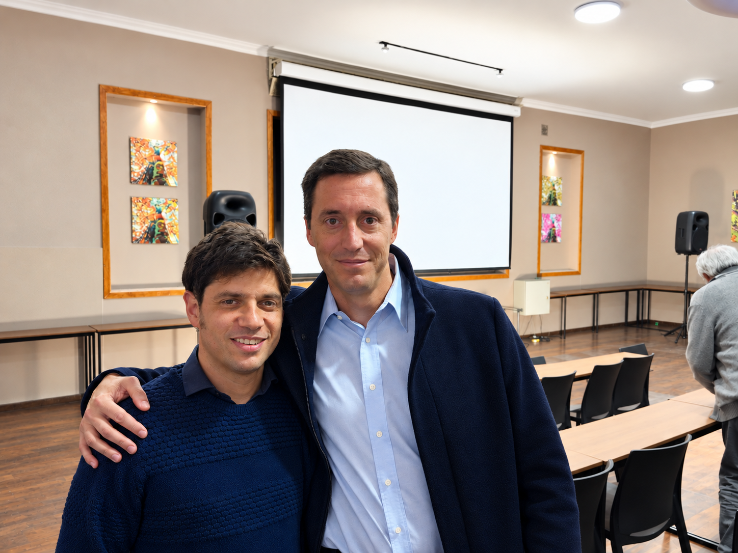 Dos personas posan abrazadas en un salón con una pantalla de proyección al fondo.